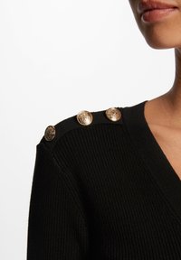 Maglione nero a coste con scollatura a V e quattro bottoni decorativi dorati sulla spalla. La texture è morbida con un design aderente.