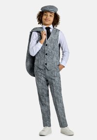 Abito grigio in tweed con gilet e berretto coordinato, camicia bianca e cravatta nera. Sneakers bianche. L'abito presenta un motivo testurizzato e quattro bottoni.