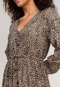 Leopardmönstrad klänning i beige och svart med V-ringning, långa ärmar, smockad midja och knapppdetalj. Slät tygtextur.