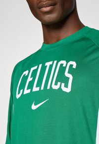 Primo piano di una persona che indossa una maglietta sportiva verde dei Celtics con un logo Nike bianco sotto il nome della squadra.