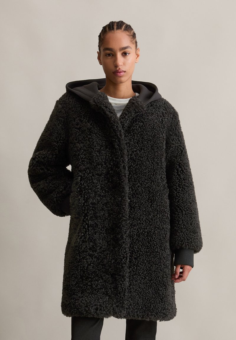 Zwarte fleece jas met een gestructureerde, fluffy buitenkant, voorzien van een grijze capuchon, lange mouwen en een rechte silhuet. Minimalistische ontwerpdetails.