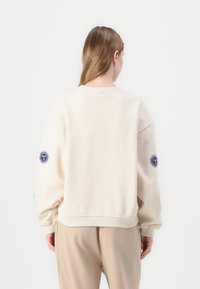 Beige sweatshirt med rundad hals och långa ärmar. Har broderade solmotiv i blått på varje ärm. Mjuk bomullsblandning.