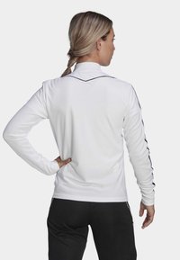 Weißes langärmliges Sportoberteil mit einem strukturierten Muster, schwarzen Akzentlinien an den Schultern und einem hohen Kragen, getragen mit schwarzen Hosen.