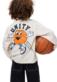 Kreem värvi pika varrukaga särk, millel on koomiksikujutis korvpallist ja seljal tekst "ÜHENDUS". Mudel hoiab käes korvpalli ja kannab musti pükse.