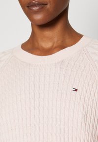 Vaaleanpunainen neulepaita, jossa on kaaplikudontakuviota, ribattu kaula-aukko ja pieni logotyyppi rinnassa.