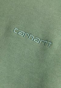 Olivgrüner Stoff mit einer strukturierten Oberfläche, versehen mit einem gestickten "carhartt"-Logo in passendem Garn, das ein dezentes Design betont.