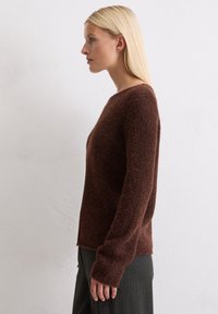 Pruun tekstuuriga kampsun ümmarguse kaelusega ja pikkade varrukatega, külgedel lõhkide ja laia lõikega. Kantud hallide pükstega.