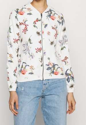 Persona che indossa una giacca bianca con zip con stampa floreale colorata abbinata a jeans azzurri chiari.
