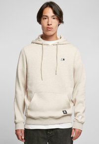 Beige hettegenser med frontlomme, snøring på hetta, ribbestrikkede mansjetter og en liten svart logo på brystet. Myk tekstur, uformelt design.