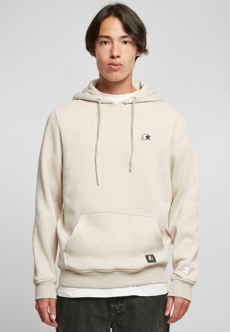 Sudadera beige con bolsillo frontal, capucha con cordón, puños acanalados y un pequeño logo negro en el pecho. Textura suave, diseño informal.
