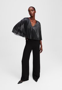 Black pleated top with a V-neckline, featuring sheer, layered sleeves. Paired with high-waisted black trousers and pointed black shoes.