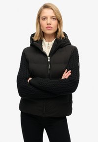 Zwarte pufferjack met een ritssluiting aan de voorkant, getextureerde kabelbreisels aan de mouwen, een verstelbare capuchon en een logo-opdruk op de linkerarm.