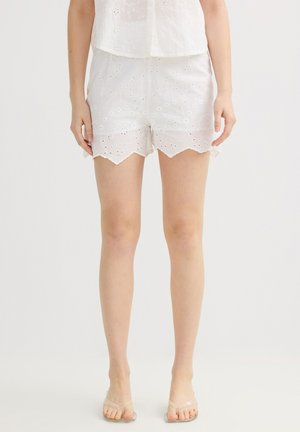 Mujer lleva pantalones cortos de encaje blanco con bajo festoneado y sandalias con tiras transparentes sobre fondo blanco liso.