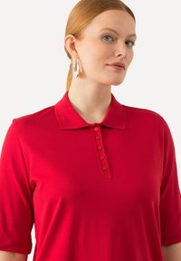 Camisa polo roja con cuello, mangas cortas y una tapeta de cinco botones. La tela parece suave, sin patrones ni acentos visibles.