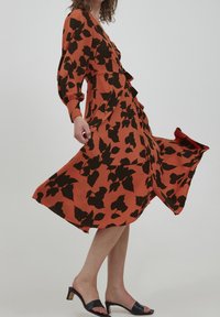 Une robe orange avec des motifs de feuilles noirs, dotée de manches longues, d'un devant volanté et d'une jupe fluide, assortie à des mules noires à talons.