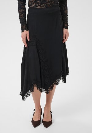Frau trägt einen schwarzen Spitzen-Midirock mit asymmetrischem Saum, ein schwarzes Spitzentop mit langen Ärmeln und spitze schwarze High Heels.