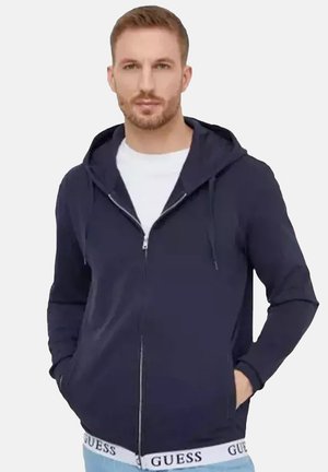 Homme portant un sweat à capuche bleu marine zippé sur une chemise blanche et un jean bleu clair avec une ceinture à logo GUESS, mains dans les poches, face à l’avant.