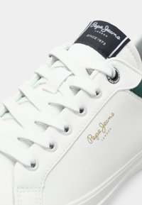 Zapatillas de cuero blanco con acentos verdes, con cordones planos, ojales metálicos y un logo en la lengüeta. Textura suave con marca en dorado.