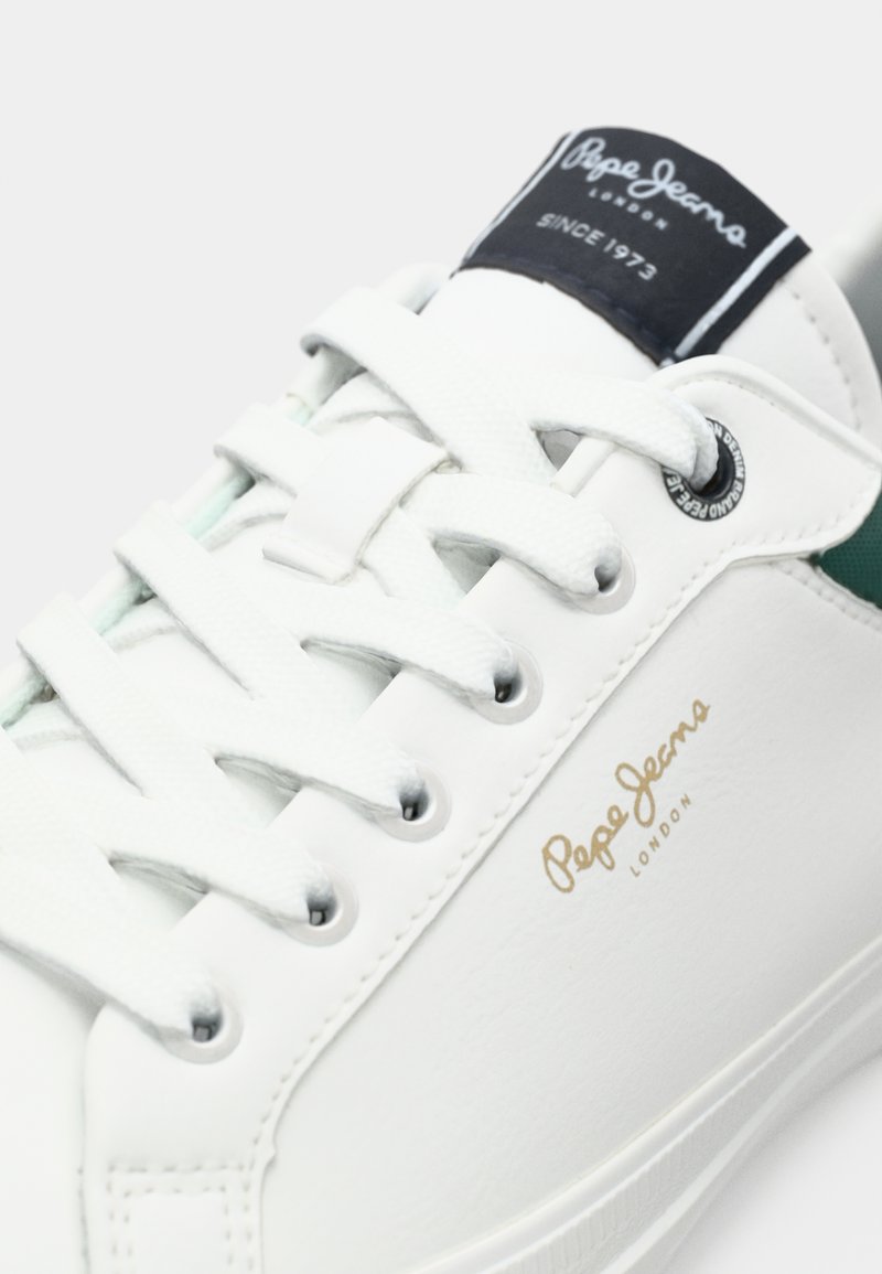 Witte leren sneaker met groene accenten, met platte veters, metalen oogjes en een logo op de tong. Soepele textuur met gouden branding.