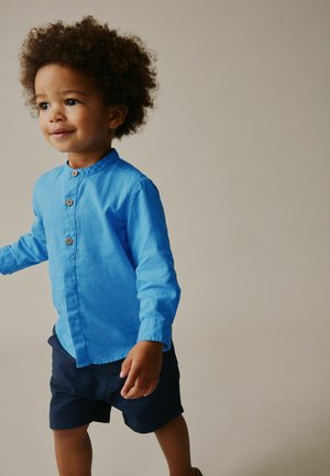 Småbarn med krøllet hår iført en klar blå skjorte med knapper og mørkeblå shorts, stående mod en ensartet beige baggrund.