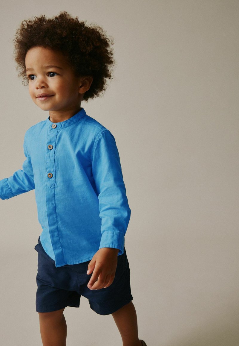 Niño pequeño con cabello rizado, vestido con una camisa azul brillante de botones y pantalones cortos azul marino, de pie frente a un fondo beige liso.