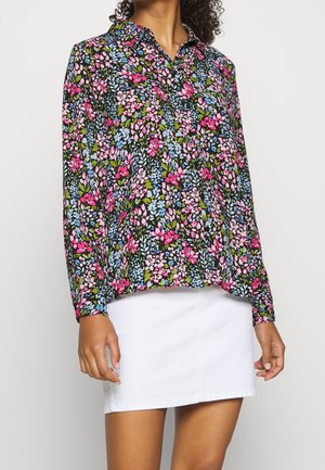 Vrouw draagt een bloemenblouse met lange mouwen met roze, blauwe en groene bloemen, gecombineerd met een witte minirok.