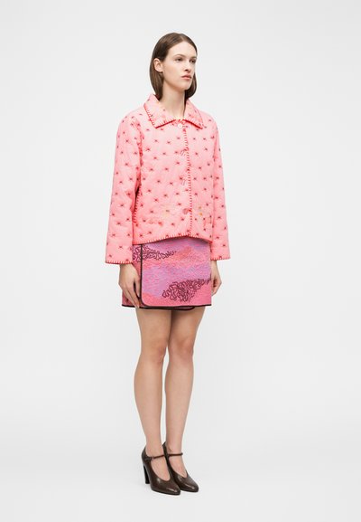 Γυναίκα που φοράει ροζ σακάκι με κουμπιά και κόκκινα μοτίβα αστεριών, ροζ υφασμένη μίνι φούστα με μαύρο περίγραμμα και σκούρα καφέ παπούτσια με λουράκι τύπου T.