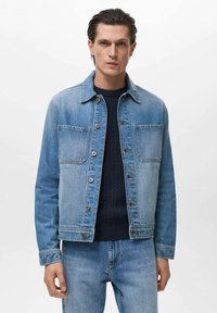 Jachetă din denim bleu deschis cu guler, închidere cu nasturi, două buzunare pe piept și o croială lejeră, purtată cu un pulover texturat bleumarin și blugi asortați.