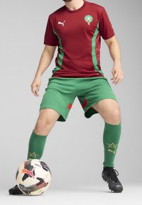 Burgundinen jalkapallo-asu vihreillä yksityiskohdilla, jossa on tunnus, yhdistetty vihreisiin shortseihin ja polveen asti yltäviin vihreisiin sukkiin, joissa on kultaiset yksityiskohdat. Mustat nappulakengät.