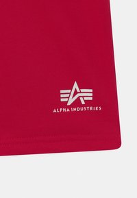 Czerwona bawełniana koszulka z srebrnym logo złożonym ze stylizowanej litery "A" i trzech równoległych linii, umieszczonym przy brzegu. Gładka faktura.