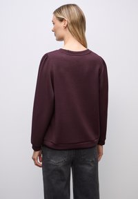 Burgundy huppari, jossa on pyöreä pääntie, pitkät hihat ja lievä puffiysitys. Yhdistetty harmaisiin housuihin, neutraalia taustaa vasten.