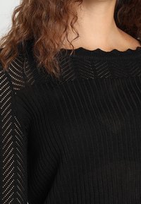 Kobieta wearing a black knit sweater with ribbed texture, openwork patterns on sleeves and yoke, and scalloped neckline detail.