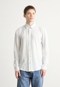 Camisa de manga longa de cor clara com um padrão xadrez subtil, colarinho clássico, fecho frontal com botões e um corte relaxado. Combinada com jeans azuis.