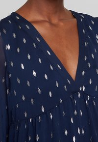 Marineblauwe jurk met een V-hals, versierd met zilverkleurige metalen stippen en een vloeiend ontwerp van zachte, gestructureerde stof.