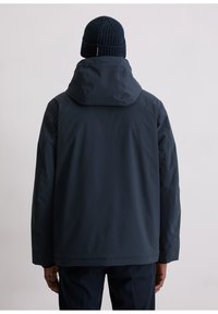 Donkerblauwe waterdichte jas met capuchon, met een gladde textuur en een rechte snit. Gebreide marineblauwe muts voegt contrast toe aan de outfit.