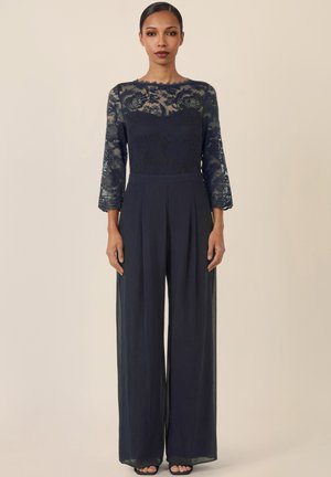 Frau trägt ein schwarzes Spitzenoberteil mit langen Ärmeln, weite schwarze Hosen und offene Schuhe, steht vor einem einfarbigen beigen Hintergrund.