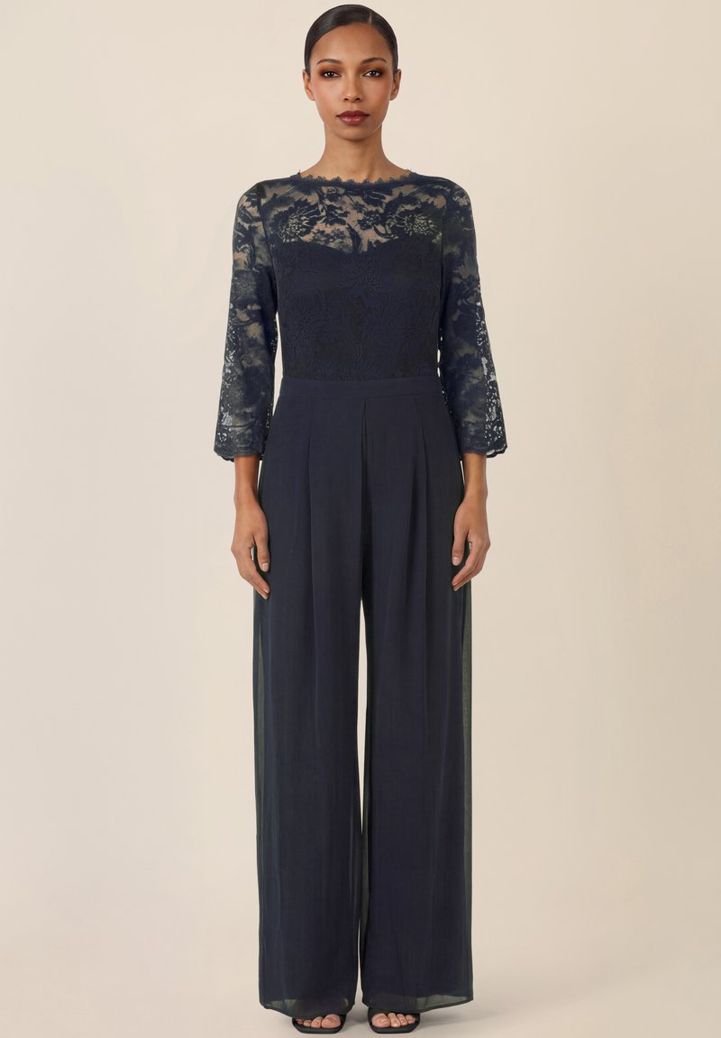 Kobieta ubrana w czarną koronkową bluzkę z długim rękawem, szerokie czarne spodnie i buty na otwartym palcu, stojąca na tle jednolitego beżowego tła.