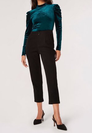 Femme portant un haut en velours bleu sarcelle à manches longues avec des épaules froncées, un pantalon court noir taille haute et des escarpins noirs pointus.