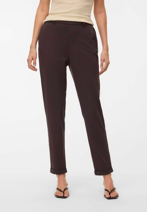Femme portant un pantalon droit marron foncé avec ourlets retroussés, un haut beige sans manches et des sandales noires à bouts ouverts, debout sur fond blanc.