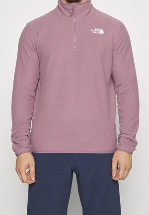 Mężczyzna ubrany w mauve polar z półzamkiem i logo The North Face na piersi oraz granatowe spodnie, stojący na jednolitym tle.