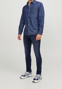 Denimskjorta i mörkblått med knäppning fram och bröstficka, kombinerad med mörka slim-fit jeans och mångfärgade sportskor.