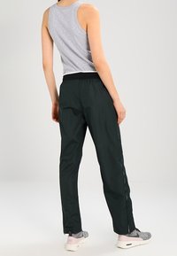 Person wearing a gray sleeveless top, black athletic pants, and gray-pink sneakers standing with back to the camera on a plain background.