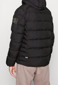 Černá péřová bunda s kapucí, s vyčiněnými horizontálními komorami a horní kapsou. Měkká, izolační tkanina a hladká textura.