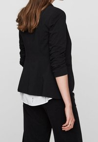 Zwart, op maat gemaakte blazer met opgerolde mouwen, gedragen over een wit overhemd. De blazer heeft een gladde textuur, een aansluitende pasvorm en subtiele details.