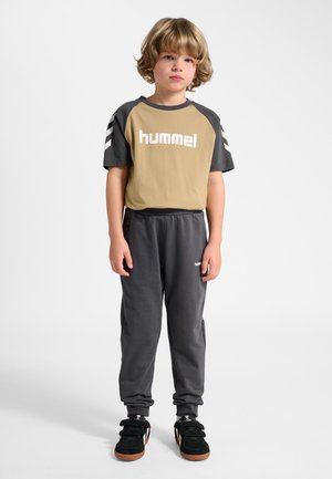 Jong kind met blond haar, gekleed in een beige en grijze hummel t-shirt, grijze joggers en zwarte sneakers, staand tegen een effen witte achtergrond.