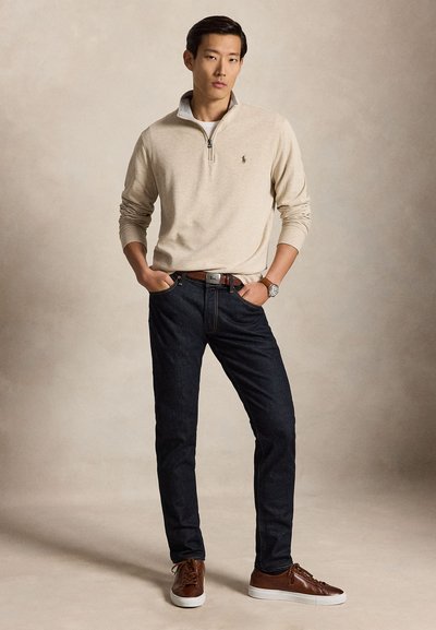 Man staat met handen in de zakken, draagt een beige trui met kwart-ritssluiting, donkere spijkerbroek, bruine riem, bruine sneakers en een polshorloge op een neutrale achtergrond.