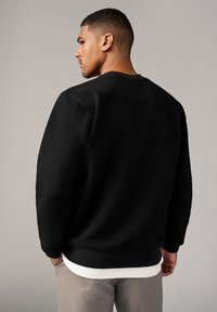 Felpa nera con texture, scollo a girocollo, polsini e orlo a coste. Indossata sopra una camicia bianca, vista da dietro. Sfondo grigio chiaro.