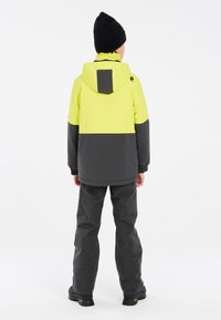 Veste jaune et grise avec capuche, dotée de poches latérales et de poignets élastiques, associée à un pantalon gris et à des chaussures noires.