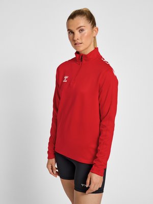 Femme portant un haut de sport rouge à manches longues zippé et un short de sport noir, debout devant un fond gris uni.