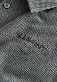 Sivá látka košele s golierom s malým károvaným vzorom, čiernym gombíkom a čiernou potlačou značky "AllSaints" na hrudi.